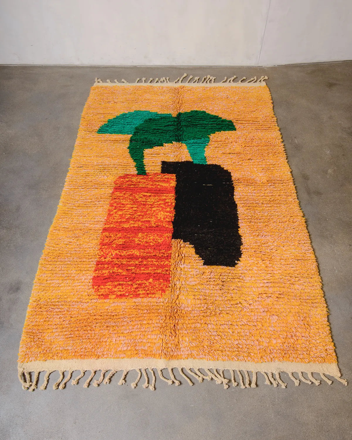 Berberlin vintage Berber rug made of wool in a background shade of mottled pink and orange, with overlapping forms in the centre in orange, black and green. The handwoven rug is laying flat on a concrete floor.